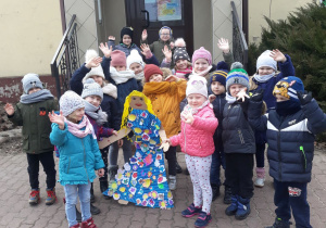 Na zdjęciu widać grupę przedszkolaków stojących przed szkołą. Każde z nich ma na sobie zimowe ubrania. Dzieci trzymają przed sobą Marzannę – czyli kukłę wykonaną z papieru oraz kolorowej bibuły, która ma na sobie niebieską sukienkę w kolorowe kwiatki. Wszyscy mają uśmiechnięte buzie.