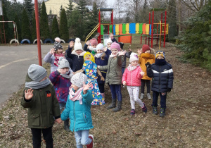 Obrazek przedstawia grupę dzieci znajdujących się na placu szkolnym. Pomiędzy dziećmi znajduje się Marzanna- czyli kukła papierowa. Wszyscy mają uśmiechnięte buzie oraz uniesione ręce ku górze. Za dziećmi znajduje się plac zabaw.
