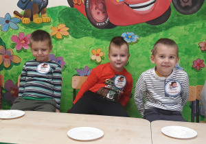 Na zdjęciu widać pięciu chłopców siedzących przy stolikach. Przed nimi znajdują się papierowe talerzyki z bitą śmietaną. Zadaniem chłopców było zjedzenie śmietany z talerza bez użycia rąk. Chłopcy mają buzie w śmietanie. Za chłopcami jest kolorowa ściana.