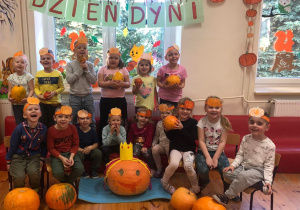 Na zdjęciu widać grupę dzieci. Każde z nich ma na głowie opaskę z dynią, wykonaną z papieru. Niektóre dzieci trzymają w rękach dynie różnej wielkości. Przed dziećmi na samym środku leży największa dynia w koronie. Wszyscy są zadowoleni.