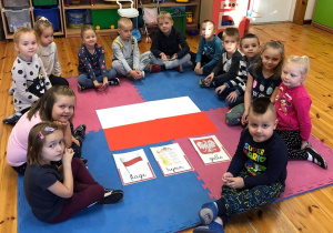 Zdjęcie przedstawia dzieci siedzące w kółku na dywanie. Pośrodku leży flaga Polski, hymn oraz godło.