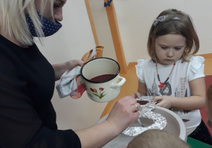 Na obrazku po lewej stronie widać Panią w czarnej sukience i jasnych włosach. Pani trzyma jasny garnek z czerwonym woskiem. Obok Pani stoi troje dzieci. Jedna dziewczynka i dwóch chłopców. Dzieci stoją wokół stolika , na którym jest miska z wodą . Dziewczynka nad miską trzyma duży, papierowy klucz, przez który Pani przelewa wosk. W tle widać papierowe nietoperze zawieszone nad drzwiami do sali.