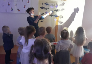 Na pierwszym planie widać grupę dzieci zwróconą twarzami do ściany. Za dziećmi stoi Pani w ciemnej sukience i w upiętych ciemnych włosach. Pani trzyma w jednej ręce telefon , którym świeci na ścianę , a w drugiej zastygniętą figurkę z wosku. Dzieci obserwują odbicie woskowej figurki na ścianie. W tle widać papierowe nietoperze, wiszące nad drzwiami do sali.