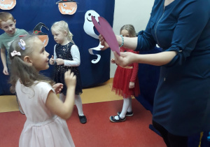 Na zdjęciu widać Panią i dziewczynkę , które są zwrócone twarzami do siebie. Pani stoi po prawej stronie i trzyma w ręku różowe serce. Ubrana jest w ciemną sukienkę i czarne buty. Dziewczynka stoi po lewej stronie i patrzy na serduszko. Ubrana jest w jasną sukienkę, rajstopy i buty. Z tyłu widać troje dzieci stojących pod ścianą i przyglądających się tej sytuacji. Wszyscy mają piękne odświętne stroje. Na ścianie jest granatowy materiał , na którym wiszą pomarańczowe dynie w czarnych kapeluszach.