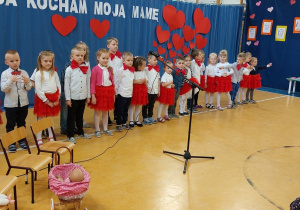 Na zdjęciu widać dzieci, które stoją na dużej sali. Przed dziećmi jest mikrofon. W tle widać dekorację z czerwonych serc.