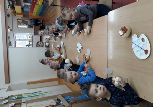 Na zdjęciu widać dzieci siedzące przy stolikach. Każde dziecko ma w rękach przygotowaną ze zgniecionego papieru kulę oraz pędzelek. Dzieci malują papier na kolor brązowy, tak aby przypominały pączki.