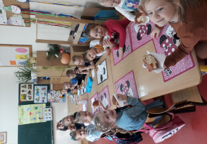 Na zdjęciu widać grupę dzieci, siedzących przy stolikach. Dzieci przed sobą mają kolorowe podkładki do jedzenia. Każde dziecko trzyma w ręce pączka owiniętego w serwetkę. W oddali widać tablicę, na której wiszą kolorowe plansze. Po prawej stronie zdjęcia są półki, na których stoją kwiaty.