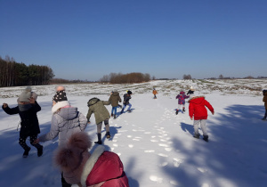 Na zdjęciu widać grupę dzieci, które chodzą po polu pokrytym śniegiem i obserwują ślady zwierząt. Dzieci maja na sobie zimowe ubrania. W oddali widać drzewa. Jest piękna , słoneczna pogoda.