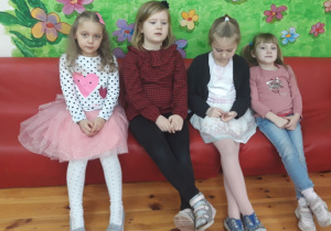 Na zdjęciu widać cztery dziewczynki siedzące na czerwonych kanapach. Każda z nich jest pięknie ubrana. Za dziećmi widać namalowaną na ścianie zieloną trawę z kwiatami.