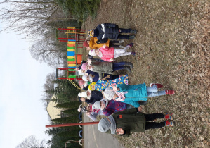 Obrazek przedstawia grupę dzieci znajdujących się na placu szkolnym. Pomiędzy dziećmi znajduje się Marzanna- czyli kukła papierowa. Wszyscy mają uśmiechnięte buzie oraz uniesione ręce ku górze. Za dziećmi znajduje się plac zabaw.
