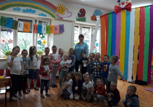 Zdjęcie przedstawia grupę dzieci wraz z Panią znajdujących się w sali przedszkola. Po prawej stronie jest duży klaun wykonany z kolorowej bibuły, zawieszony przy suficie. Za dziećmi jest napis Dzień Dziecka. Wszystkie dzieci są zadowolone.