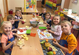 Na obrazku widać dzieci siedzące przy stolikach. Każde z nich ma przed sobą deskę do krojenia, na której leżą różne rodzaje owoców. Dzieci kroją owoce, przygotowując sałatkę owocową. Wszyscy są zadowoleni i uśmiechnięci.