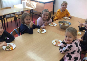 Na zdjęciu widać dzieci siedzące przy stolikach. Przed sobą mają talerzyki, na których jest różnokolorowa sałatka owocowa. Na środku stołu jest miska z sałatką. Dzieci jedzą przygotowaną wcześniej przez siebie sałatkę.
