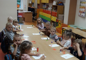 Na zdjęciu widać kilkoro dzieci siedzących przy stolikach. Dzieci mają przy sobie przybory plastyczne oraz kartki, na których widnieją sygnalizatory świetlne. Zadaniem dzieci jest wyklejenie kółek sygnalizatora odpowiednimi kolorami.