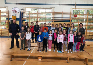 Zdjęcie przedstawia grupę dzieci, które stoją na sali gimnastycznej. Dzieci ustawione są w dwóch rzędach. Obok dzieci stoją Panie oraz Pan Policjant.