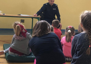 Za zdjęciu widać kilkoro dzieci siedzących na materacach. Na drugim planie widać policjanta siedzącego przy stoliku, wygłaszającego pogadankę. Dzieci są skupione i uważnie słuchają.