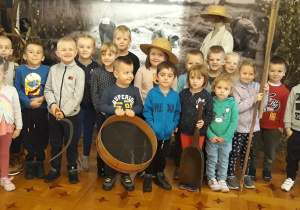 Na zdjęciu widać grupę dzieci, które znajdują się w pomieszczeniu o aranżacji przedstawiającej pracę chłopów na wsi podczas żniw. Kilkoro dzieci wyposażonych jest w rekwizyty typu: kapelusz, sierp, cepy, szufla oraz przetak.