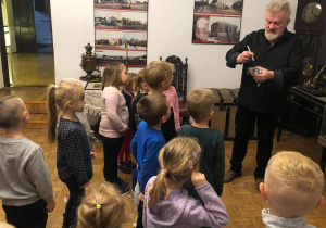 Na obrazku widać grupę dzieci znajdujących się w muzeum. Obok nich po prawej stronie stoi przewodnik, który opowiada jak wygląda kałamarz. Wszystkie dzieci wyglądają na zaciekawione. Na drugim planie, na ścianie widać zdjęcia, przedstawiające jak kiedyś wyglądało miasto.