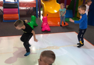 Na obrazku widać kilkoro dzieci bawiących się w sali zabaw. Dzieci grają na dywanie interaktywnym. Z tyłu widać zjeżdżalnie, trampolinę oraz tor przeszkód.