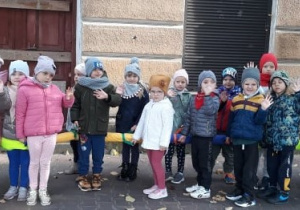 Na zdjęciu widać grupę dzieci stojących na chodniku. Po prawej stronie stoi Pani. Każde z nich ubrane jest w ciepłe ubrania i czapki, dzieci machają rękami. Za dziećmi znajduje się budynek.