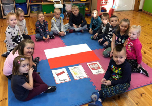 Zdjęcie przedstawia dzieci siedzące w kółku na dywanie. Pośrodku leży flaga Polski, hymn oraz godło.