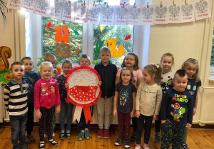 Na zdjęciu widać grupę dzieci, jedno z nich trzyma w rękach piękny, duży kotylion w kolorach narodowych. Nad dziećmi są obrazki przedstawiające godło Polski.