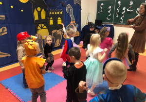 Na zdjęciu widać grupę dzieci, które naśladują Panią przedstawiająca jakiś taniec. Dzieci mają na sobie stroje andrzejkowe. Na drugim planie widać dekorację przedstawiającą zamek, nietoperze oraz duchy.