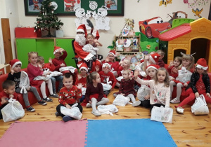 Na zdjęciu widać Św. Mikołaja oraz dzieci siedzące wokół niego. Każde trzyma w ręku prezent. Wszyscy są uśmiechnięci i zadowoleni