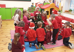 Zdjęcie przedstawia grupę dzieci, które ubierają drzewko świąteczne. Każde z nich ma na sobie ubranie w czerwonym kolorze.