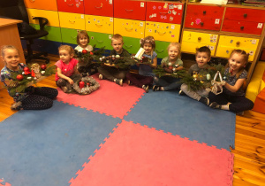 Na zdjęciu widać grupę dzieci, które siedzą na dywanie. Każde z nich trzyma przed sobą własnoręcznie wykonany stroik bożonarodzeniowy.