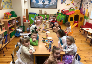 Na zdjęciu widać dzieci siedzące przy stolikach pełnych różnych smakołyków. Dzieci mają piękne, kolorowe stroje. Każdy jest uśmiechnięty.