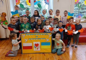 Na zdjęciu widać grupę dzieci. Część z nich trzyma łańcuszek z biało- czerwonych serc. Niektórzy mają w rękach motylki w kolorach flagi Ukrainy. Przed dziećmi znajduje się gazetka, na której są elementy symbolizujące Solidarność z Ukrainą.