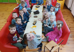 Na zdjęciu widać grupę dzieci, które siedzą na czerwonych kanapach. Pomiędzy nimi stoją stoliki ze słodkim poczęstunkiem.