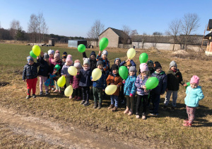 Dzieci z oddziału przedszkolnego stoją na łące. Każde z nich trzyma zielony balon. Jest piękna, słoneczna pogoda.