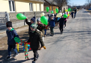Zdjęcie przedstawia grupę dzieci, które spacerują po okolicy z Marzanną. Każde z nich trzyma w ręku zielony balon.