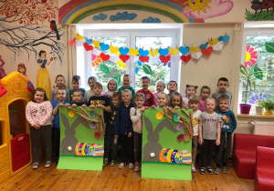 Na zdjęciu widać grupę dzieci. Dzieci trzymają w rękach dwa identyczne plakaty, przedstawiające zająca na zielonym obok niego są kolorowe pisanki.