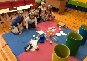 Dzieci siedzą na dywanie pełnym różnych śmieci. Każde z nich ma na głowie opaskę z symbolem ziemi.