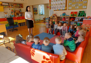 Zdjęcie przedstawia Panią oraz grupę dzieci, które siedzą na czerwonych kanapach w sali przedszkolnej.