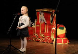 Na zdjęciu widać dziewczynkę stojącą przy mikrofonie, za nią stoi czerwona lokomotywa.