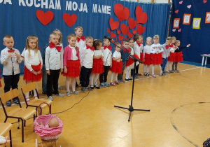 Na zdjęciu widać dzieci, które stoją na dużej sali. Przed dziećmi jest mikrofon. W tle widać dekorację z czerwonych serc.