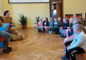 Zdjęcie przedstawia dwie Panie, naprzeciwko których siedzi grupa dzieci. Wszyscy znajdują się w dużej sali.