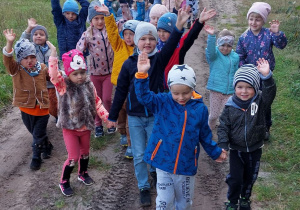 Grupa dzieci wraz z Panią idzie na spacer polną drogą w kierunku lasu.